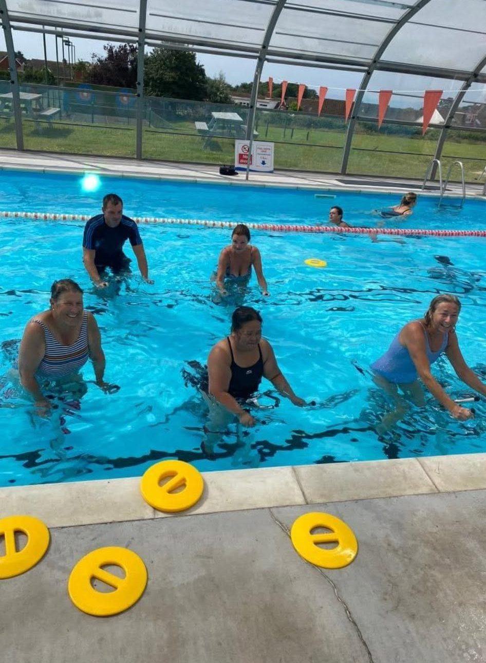 A sunny outdoor Aquaspin class with smiling riders on bikes in the pool and countryside behind